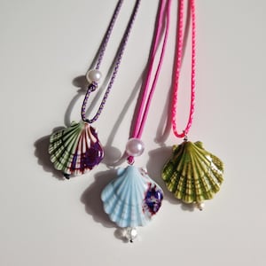 Peut inclure: Trois colliers avec des pendentifs en forme de coquillage. Les colliers sont faits de cordons de différentes couleurs : violet, rose et rose vif. Chaque pendentif est d’une couleur différente : vert et violet, bleu et violet, et vert.