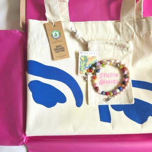 May include: Cream-colored tote bag with blue abstract design and a tag that reads "100% Organic Cotton." A small drawstring bag with a colorful beaded bracelet and the text "STUDIO AMMOS" is also visible.