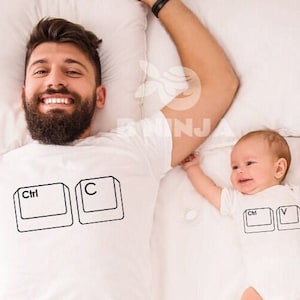Puede incluir: Una camiseta blanca con un gráfico negro de teclas de computadora que dicen "Ctrl" y "C". Un bebé que lleva un mameluco blanco con un gráfico negro de teclas de computadora que dicen "Ctrl" y "V" está acostado en una cama blanca.