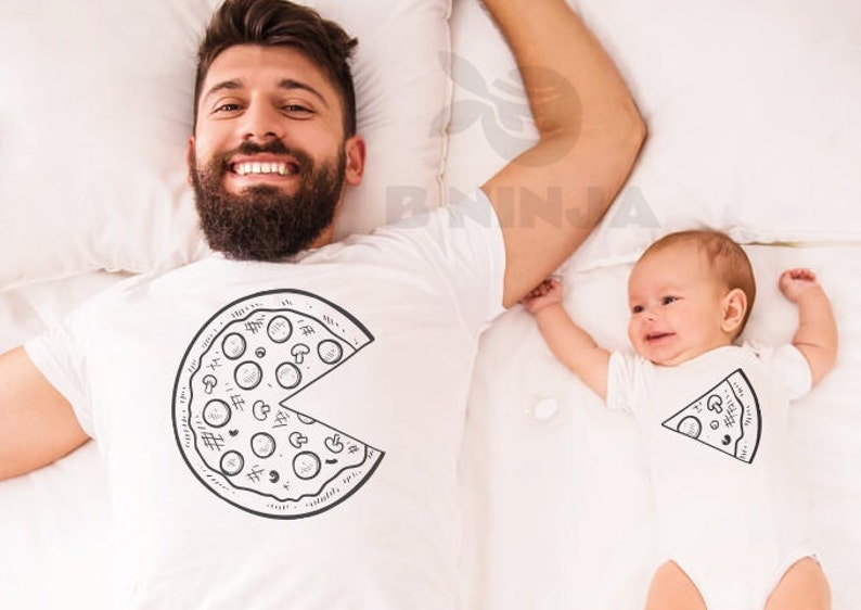 May include: A father and child wearing matching white t-shirts with a pizza graphic. The father's shirt has a whole pizza with a slice missing, while the child's shirt has a single slice of pizza.