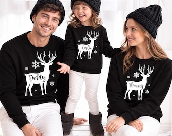 May include: Three black long-sleeved sweatshirts featuring a white graphic of a reindeer with snowflakes. The sweatshirts are personalized with the text Daddy, Leslie, and Mommy printed on the body of each respective reindeer graphic.