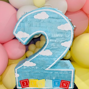 May include: A light blue number 2 piñata with white cloud cutouts and a white border. The piñata has the name "JULIEN" spelled out in colourful letter blocks. The background features pink, white, and yellow balloons, creating a festive atmosphere.