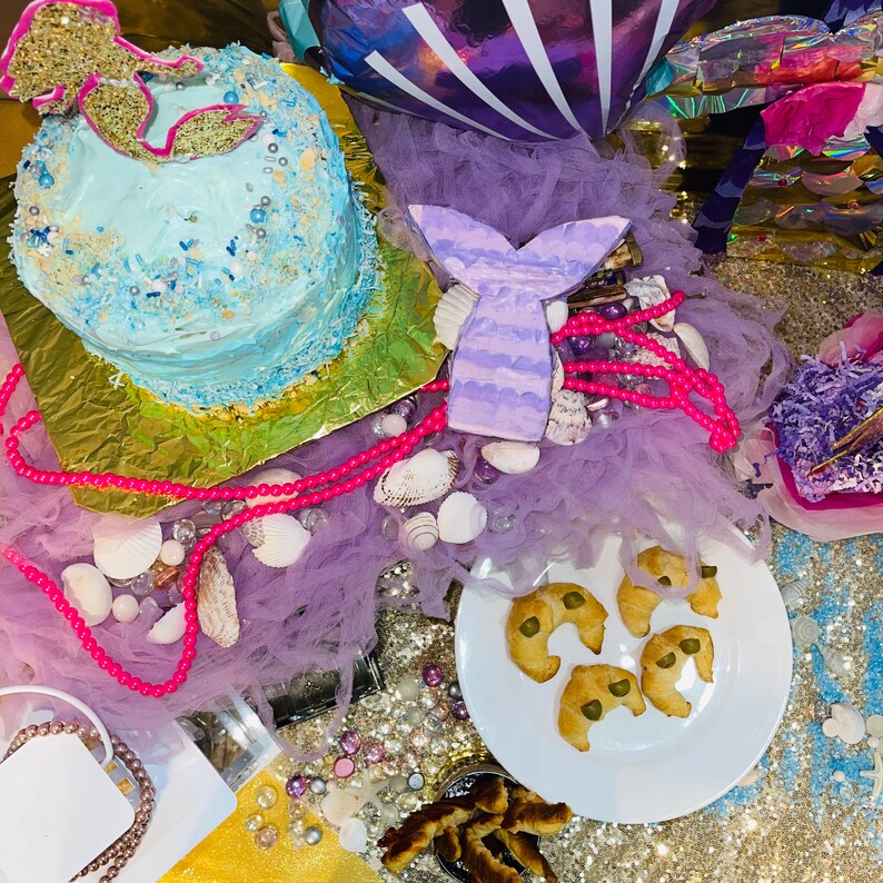 Puede incluir: Una escena de fiesta con tem&aacute;tica de sirenas con un pastel azul claro decorado con chispas y una cola de sirena dorada. Tambi&eacute;n se ven una decoraci&oacute;n de cola de sirena p&uacute;rpura, conchas marinas, cuentas rosas y galletas.