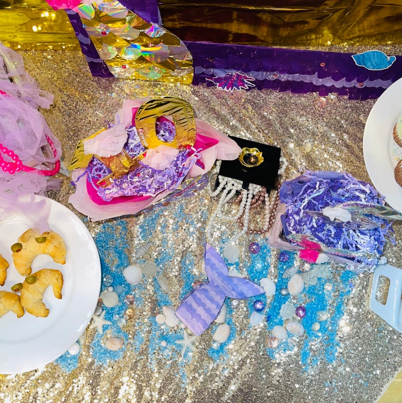 Puede incluir: Una exhibici&oacute;n con tem&aacute;tica de sirenas con un fondo dorado con lentejuelas. La escena incluye una cola de sirena de papel, joyas y un plato de comida. Los elementos decorativos incluyen conchas marinas, estrellas de mar y acentos morados y azules.