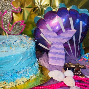 Puede incluir: Una tarta de cumplea&ntilde;os con tem&aacute;tica de sirena con glaseado azul y un adorno de tarta de sirena con purpurina. La escena incluye una decoraci&oacute;n de cola de sirena, conchas marinas y cuentas rosas.