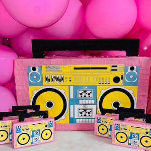 Puede incluir: Una gran piñata de boombox rosa con un asa negra y detalles amarillos, azules y blancos. Varios pequeños recuerdos de fiesta con forma de boombox rosa están frente a la piñata. El fondo está lleno de globos rosas.