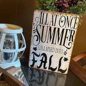 May include: White wooden sign with black lettering that reads "All at once summer collapsed into fall".