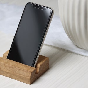 Könnte beinhalten: Ein Telefonständer aus hellem Holz mit einem Smartphone. Der Ständer hat eine Aussparung, um das Telefon aufrecht zu halten. Der Bildschirm des Telefons ist schwarz. Im Hintergrund ist eine weiße Vase zu sehen.