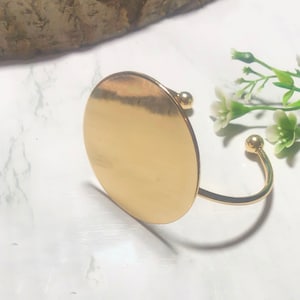 May include: A gold-toned bangle bracelet with a large, round, flat disc charm. The bracelet is open and has a simple, minimalist design.