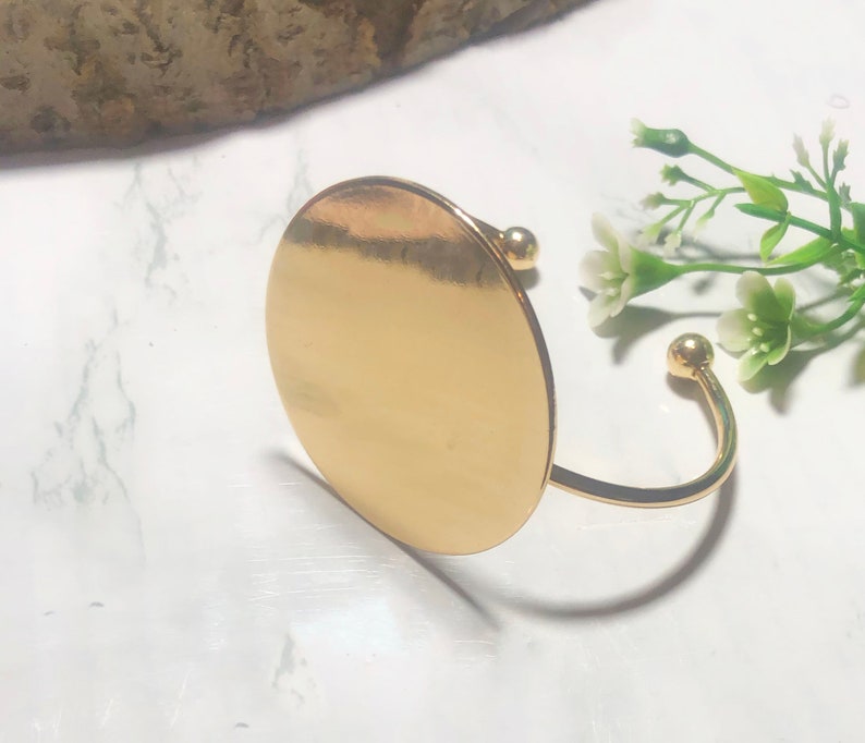 May include: A gold-toned bangle bracelet with a large, round, flat disc charm. The bracelet is open and has a simple, minimalist design.