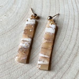 Peut inclure: Une paire de longues boucles d'oreilles rectangulaires avec un motif marbré blanc et or. Les boucles d'oreilles ont des crochets en métal doré.