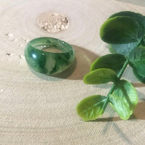 Op de afbeelding: Een groene jade ring met witte swirls. De ring ligt op een licht houten oppervlak met groene bladeren op de achtergrond.
