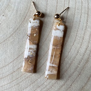 K&ouml;nnte beinhalten: Ein Paar goldene und wei&szlig;e gestreifte Ohrringe. Die Ohrringe sind aus Harz und haben eine goldfarbene Oberfl&auml;che.