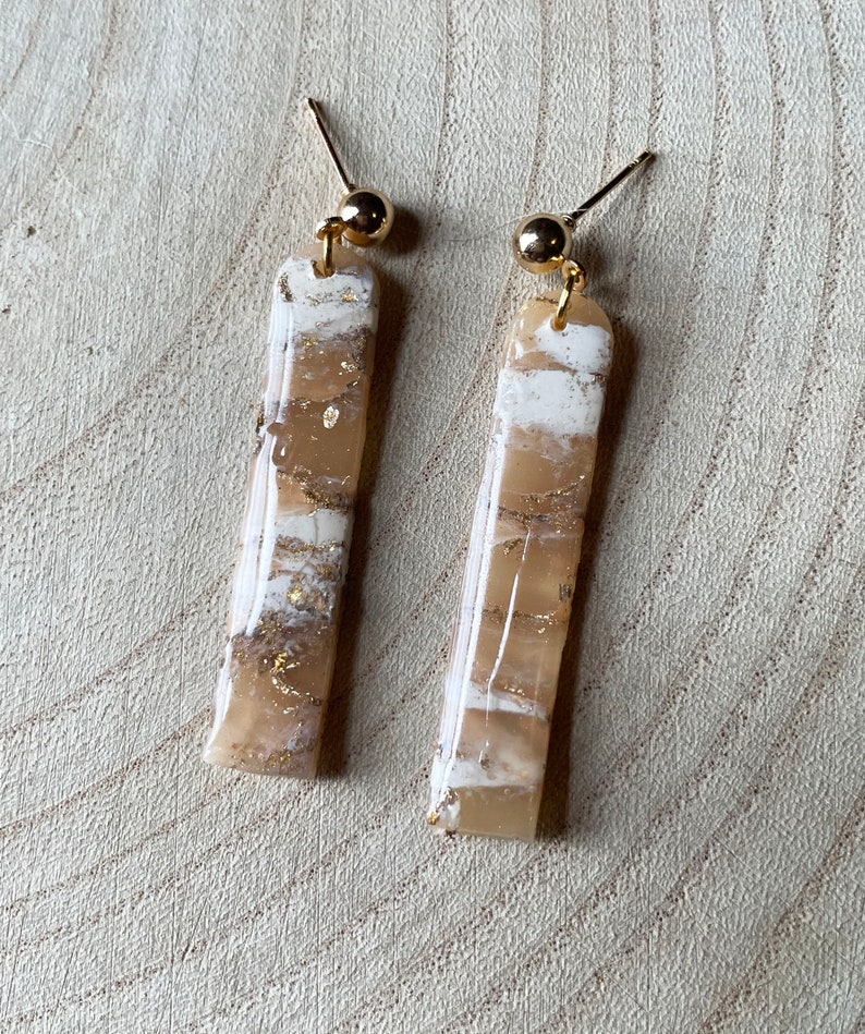 K&ouml;nnte beinhalten: Ein Paar goldene und wei&szlig;e gestreifte Ohrringe. Die Ohrringe sind aus Harz und haben eine goldfarbene Oberfl&auml;che.