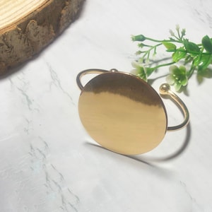 May include: A gold-toned bangle bracelet with a large, circular, flat disc. The disc is smooth and shiny.