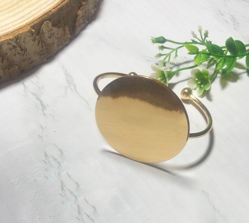 May include: A gold-toned bangle bracelet with a large, circular, flat disc. The disc is smooth and shiny.