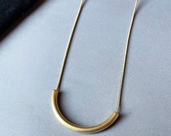Puede incluir: Un collar dorado con una cadena delgada y un colgante curvo en forma de tubo. El colgante tiene un acabado mate y está suspendido de una delicada cadena. El collar se muestra sobre una superficie gris.