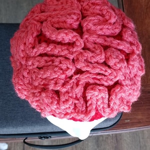 May include: A handmade, coral-colored crochet brain hat. The hat is designed to resemble a human brain, with intricate folds and ridges. The hat is sitting on a white surface.