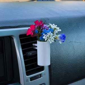 May include: A white plastic car air freshener with a bouquet of pink, blue, and white flowers. The air freshener is attached to the air vent in a car.