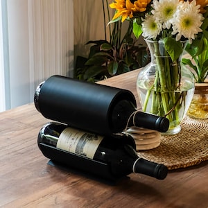 May include: A black metal wine rack holds two bottles of red wine on a wooden table. The bottles are labeled with a white label and have a black cap.