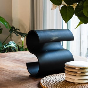 May include: Black metal wine bottle holder with a wavy design, sitting on a wooden table with a woven placemat and a stack of white coasters.