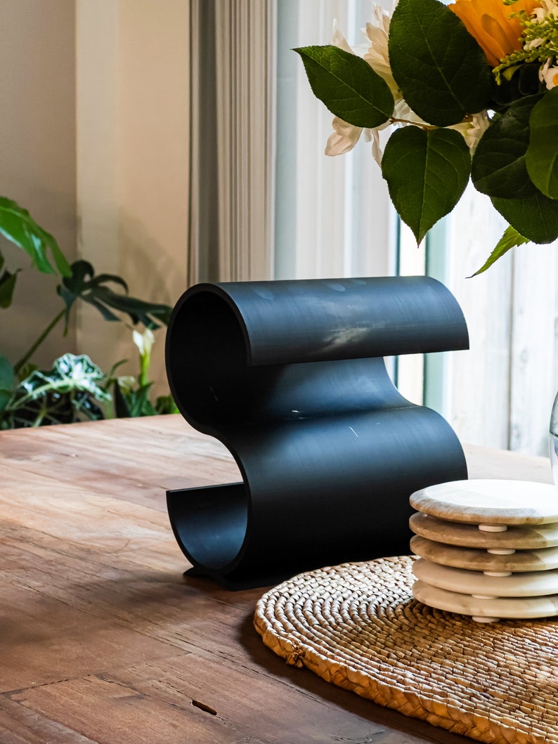 May include: Black metal wine bottle holder with a wavy design, sitting on a wooden table with a woven placemat and a stack of white coasters.