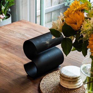 May include: A black metal wine bottle holder shaped like an 'S' on a wooden table with a woven placemat and a stack of white marble coasters.