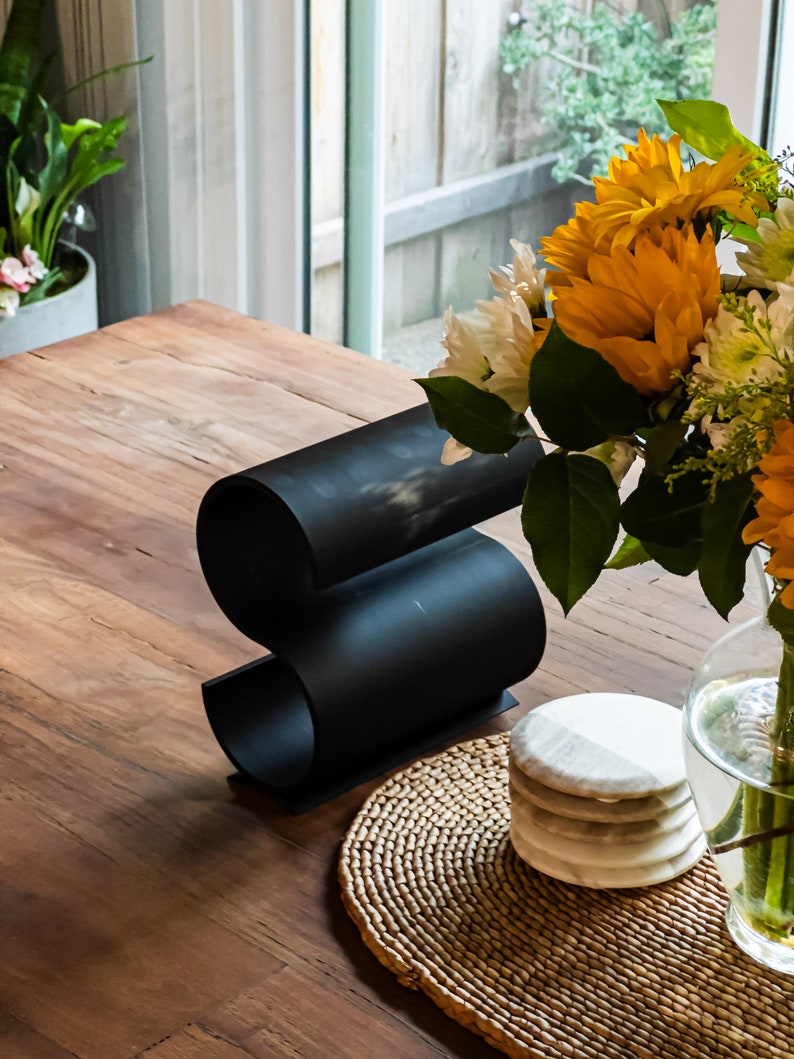 May include: A black metal wine bottle holder shaped like an 'S' on a wooden table with a woven placemat and a stack of white marble coasters.
