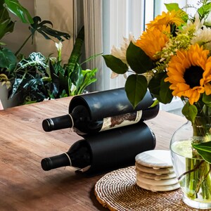 May include: A black metal wine rack with two bottles of wine. The rack is on a wooden table with a woven placemat and coasters. A bouquet of yellow and white flowers in a clear glass vase is on the table.