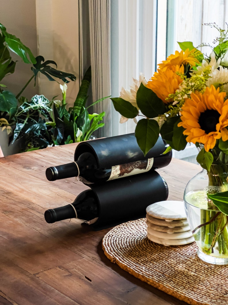 May include: A black metal wine rack with two bottles of wine. The rack is on a wooden table with a woven placemat and coasters. A bouquet of yellow and white flowers in a clear glass vase is on the table.