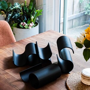 May include: Black metal wine rack with a wavy design. The rack is made of four pieces that fit together to create a unique shape. The rack is sitting on a wooden table.