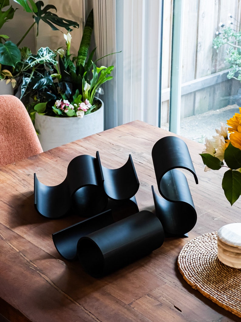 May include: Black metal wine rack with a wavy design. The rack is made of four pieces that fit together to create a unique shape. The rack is sitting on a wooden table.