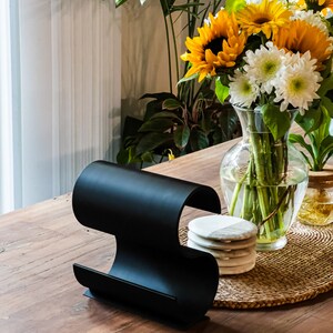 May include: A black metal magazine rack with a curved design sits on a wooden table. A vase with sunflowers and white flowers sits on a woven placemat with white coasters.