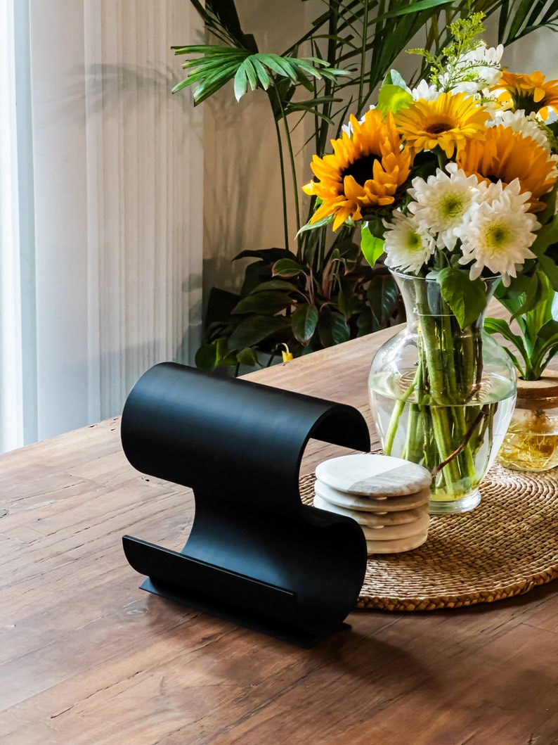 May include: A black metal magazine rack with a curved design sits on a wooden table. A vase with sunflowers and white flowers sits on a woven placemat with white coasters.