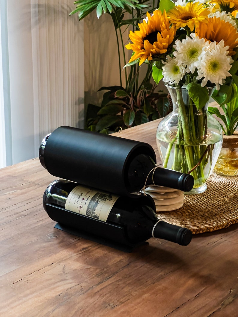 May include: A black metal wine rack holds two bottles of red wine on a wooden table. The bottles are labeled with a white label and have a black cap.