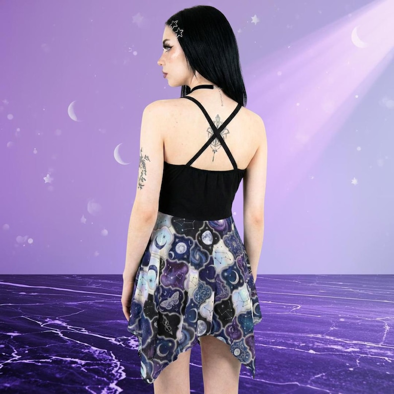 May include: A black tank top with crisscross straps and a flared skirt. The skirt features a celestial pattern with moons, stars, and constellations in shades of blue, purple, and white. The outfit is set against a purple background.