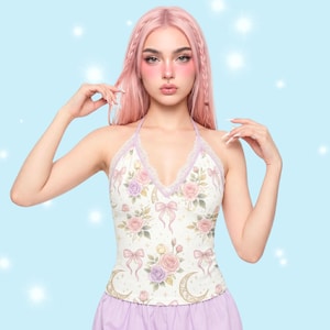 May include: A halter top with a floral and bow pattern. The top features a white base with pink and purple roses, gold crescent moons, and pink bows. The neckline is trimmed with lace and a lavender strap. The top is a light, airy style.