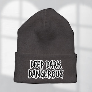 May include: A dark gray beanie with the words "DEEP DARK DANGEROUS" embroidered in white, stylized lettering. The beanie is a knit cap, suitable for cold weather.