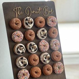 May include: A wooden donut bar display with the words "Welcome to The Donut Bar" in gold script. The display holds 20 donuts with various toppings, including sprinkles and chocolate. A plate of donuts sits below.