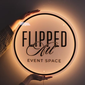 May include: A round, illuminated sign with the text "FLIPPED Atl EVENT SPACE" in black. The sign has a black border and a warm, glowing light around the edges. The sign is held up by a person.