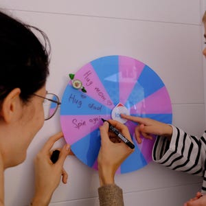 Peut inclure: Une roue tournante rose et bleue avec les mots "Hug mom", "Hug dad" et "Spin again" écrits dessus. Une jeune fille pointe la roue tandis qu'une femme écrit dessus avec un marqueur noir.