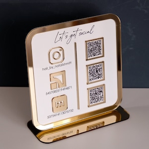 Op de afbeelding: Een goudomrand, wit acryl social media display stand. De stand heeft Instagram, een logo en QR-codes. De tekst "Let's get social" staat bovenaan. De Instagram-handle is "hair_by_natalishmitt".
