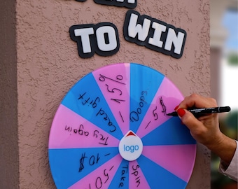 May include: A colorful prize wheel with blue and pink segments, labeled with phrases like "Spin Again" and "Take a Gift." The words "SPIN TO WIN" are displayed above the wheel. A hand is writing on the wheel with a black marker.