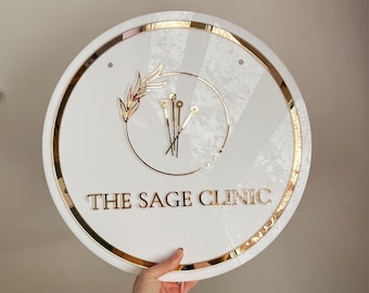 May include: A round white sign with gold trim and lettering that reads "THE SAGE CLINIC." The sign features gold leaf and line art. The sign is being held up by a person.