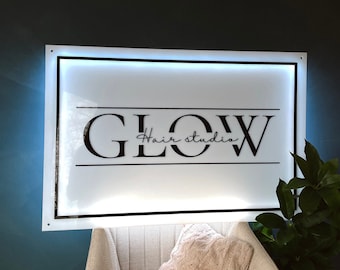 May include: A white sign with black lettering that reads "GLOW Hair Studio" with a white light behind the sign. The sign is mounted on a blue wall.