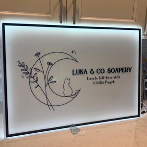 May include: A white sign with a black border features the text "LUNA & CO. SOAPERY" and "Elevate Self-Care With A Little Magick." A crescent moon, cat silhouette, and floral design are also included.