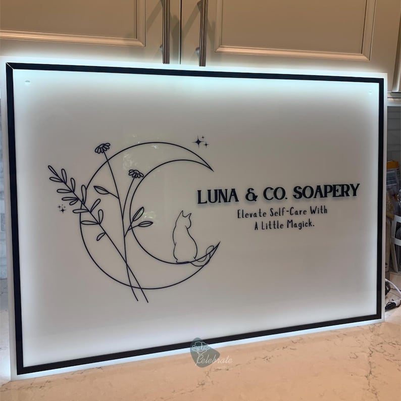 May include: A white sign with a black border features the text "LUNA & CO. SOAPERY" and "Elevate Self-Care With A Little Magick." A crescent moon, cat silhouette, and floral design are also included.