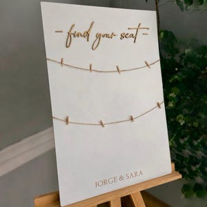 Puede incluir: Un letrero blanco con las palabras "- find your seat -" en fuente cursiva. Dos cuerdas con pequeñas pinzas para la ropa están adjuntas. Los nombres "JORGE & SARA" están impresos en la parte inferior. El letrero se muestra en un caballete de madera.