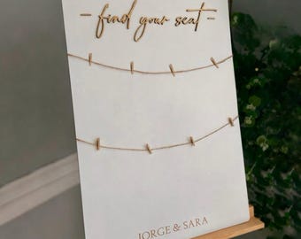 May include: A white sign with the words "- find your seat -" in a cursive font. Two strings with small clothespins are attached. The names "JORGE & SARA" are printed at the bottom. The sign is displayed on a wooden easel.
