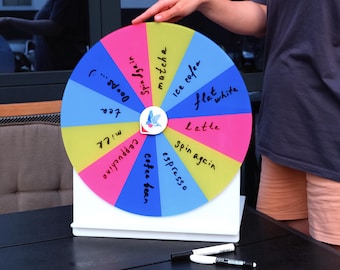 May include: A colorful prize wheel with segments labeled for coffee drinks like "latte" and "espresso." The wheel is mounted on a white stand, with markers nearby. The wheel's segments are in shades of pink, blue, and yellow.